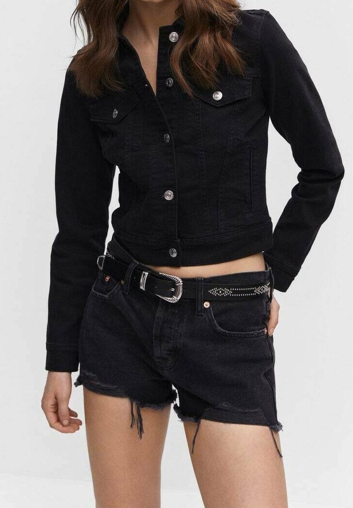 Femme portant une veste en jean noire boutonnée et un short en jean noir effiloché avec une ceinture noire cloutée sur un fond uni.
