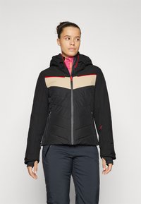 Svart isolert jakke med et beige og rødt sikk-sakk mønster, hette, glidelås foran og sidelommer, med en quiltet tekstur.
