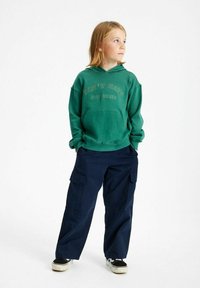 Sudadera verde con capucha con el texto "NO ODIES", combinada con pantalones cargo azul marino con bolsillos. Zapatillas negras y blancas completan el look.