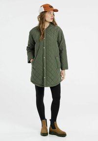 Prešívaný zelený dlhý kabát so zapínaním na patentky, bočnými vreckami, nosený cez čierne legíny a hnedé členkové čižmy, v kombinácii s oranžovou čiapkou.