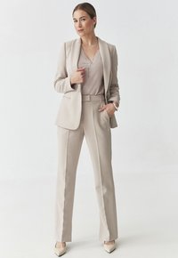 Completo beige su misura con giacca aderente e pantaloni a gamba larga. Tessuto morbido, top scollo a V e tacchi in tonalità neutra completano il look.