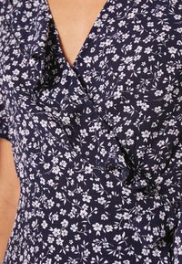 Marine dress fabric med hvit blomstermønster, med volangkant i halsen og v-hals design. Nærbilde av tekstur og detaljer.