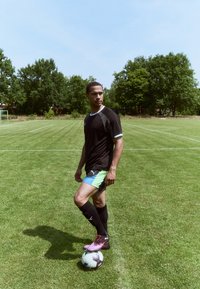 Athlète en maillot noir à manches courtes et short avec des accents verts, debout sur un ballon de football sur de l'herbe, portant des chaussettes noires et des chaussures à crampons roses.