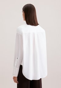Camicia bianca a maniche lunghe con orlo curvo, dettaglio plissettato sul retro e polsini con bottoni che presentano un piccolo design ricamato. Tessuto liscio.