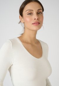 Langsleeve top in een lichte crèmekleur, zachte textuur, met een diepe V-halslijn, slanke pasvorm en subtiele stikaccenten rond de halslijn.