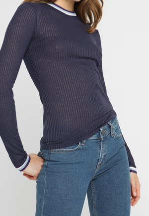 Marineblaues, ribbeltes Langarmoberteil mit transparenter Textur und kontrastierendem gestreiften Halsausschnitt. Kombiniert mit eng sitzenden blauen Jeans.