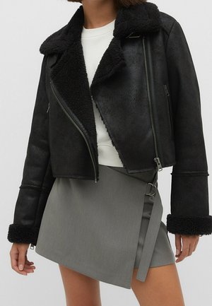 Vrouw draagt een zwarte shearling jas met ritssluitingen over een witte top en een grijze asymmetrische overslagrok met een zijriem detail.