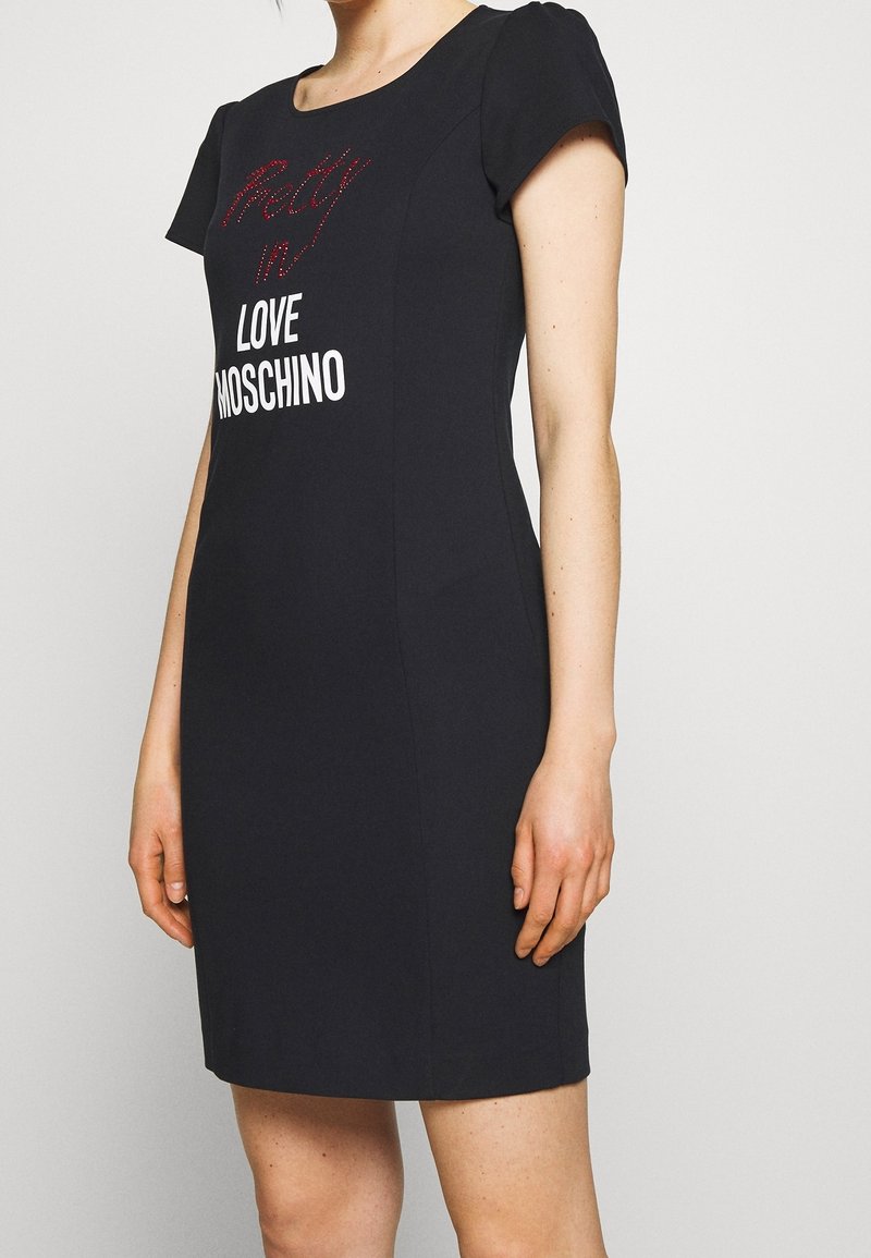 Femme portant une robe noire à manches courtes avec un texte rouge et blanc indiquant "Pretty in Love Moschino" à l'avant.