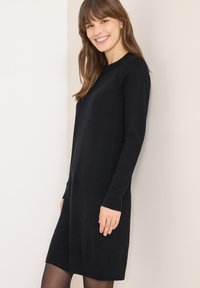 Robe en maille noire à manches longues, col rond et poignets côtelés. Arrive juste au-dessus des genoux, présente une texture lisse et un design minimaliste.
