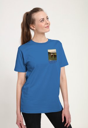 Donna con coda di cavallo lunga che indossa una maglietta blu con la riproduzione del dipinto di una corsa di cavalli di Edgar Degas sul petto, che guarda verso l'alto sorridendo leggermente.