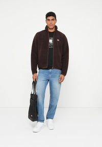 Jachetă cu fermoar din fleece maro, cu guler înalt, blugi albaștri, adidași albi și o geantă tote neagră, având accente cu marcă.