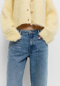 Sárga, bolyhos, rövidített kardigán gombos záródással, kék farmernadrággal párosítva, amelynek elülső részén masni alakú hímzés található.