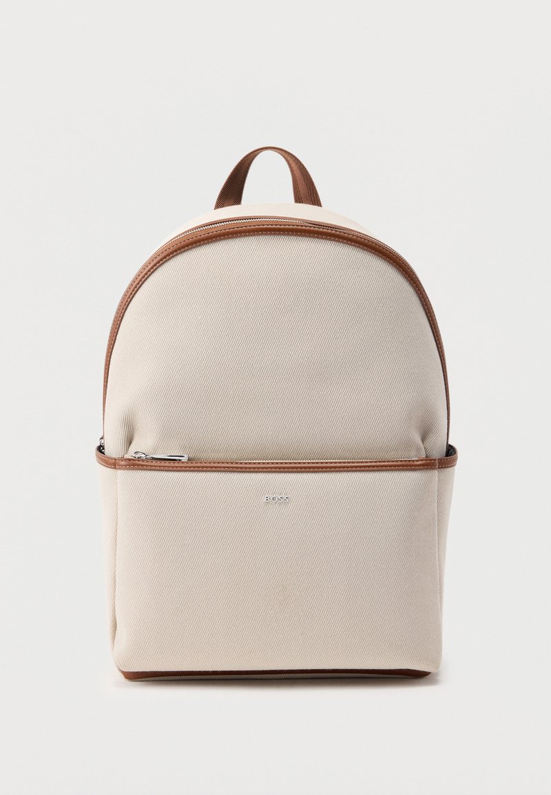 Sac à dos en tissu beige avec garniture marron, poche avant zippée, poignée supérieure et logo métallique "BOSS" sur fond blanc uni.