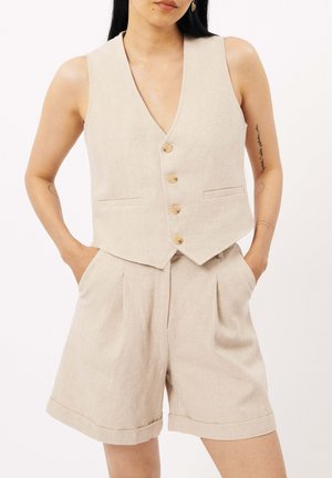 Gilet en lin beige avec un profond décolleté en V, cinq boutons en bois et deux poches avant, associé à un short taille haute assorti avec des plis.