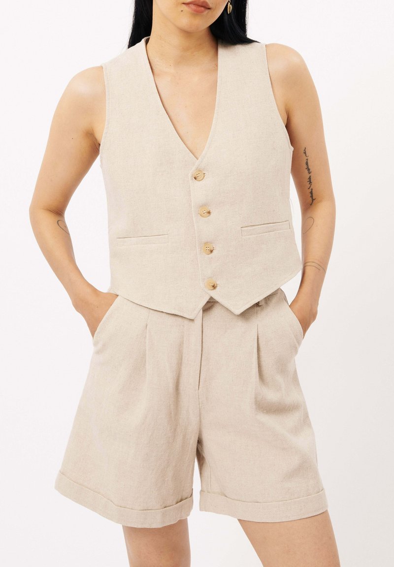 Gilet en lin beige avec un profond décolleté en V, cinq boutons en bois et deux poches avant, associé à un short taille haute assorti avec des plis.