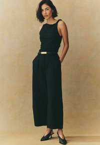 Top nero senza maniche con scollatura ondulata, abbinato a pantaloni neri a vita alta e gamba larga e tacchi slingback neri.