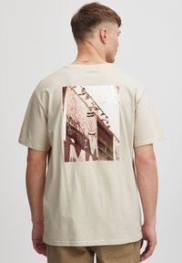Beige bomulds t-shirt med et stort grafisk print på ryggen, der fremstiller sneakers på en væg dækket af graffiti. Korte ærmer, afslappet pasform.