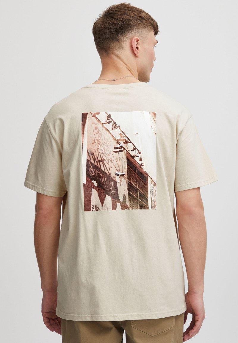 Beige bomulds t-shirt med et stort grafisk print på ryggen, der fremstiller sneakers på en væg dækket af graffiti. Korte ærmer, afslappet pasform.