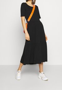Robe maxi noire à manches courtes, design à étages, tissu fluide, portée avec des baskets blanches et un sac bandoulière orange.
