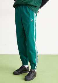 Zelené sportovní kalhoty s bílými bočními pruhy, elastickým pasem a pružnými manžetami, doplněné černými teniskami.