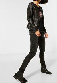 Černá koženka s kulatým výstřihem, doplněná černým tričkem s grafikou a tmavými skinny džíny. Obuv zahrnuje černé boty do kotníků.