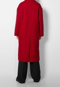 Abrigo de lana rojo con corte recto, mangas largas, cuello de muesca y abertura en la parte posterior. Combinado con pantalones sueltos negros.