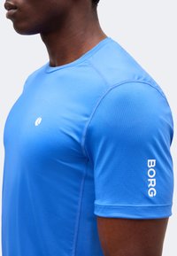 Άνδρας που φοράει φωτεινό μπλε αθλητικό κοντομάνικο μπλουζάκι με το κείμενο «BORG» στο μανίκι και ένα μικρό λευκό λογότυπο στο στήθος.