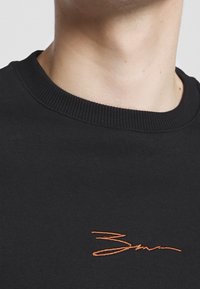 Primo piano del collo e della parte superiore del torace di una persona che indossa una maglietta nera a girocollo con un piccolo testo ricamato arancione.