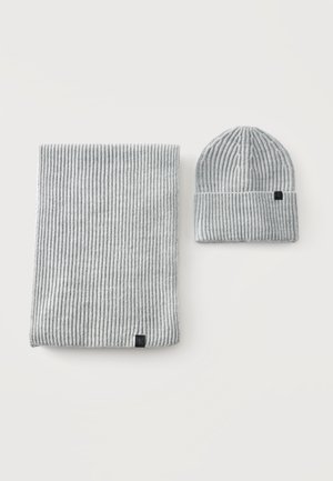 Écharpe tricotée côtelée gris clair et bonnet assorti, tous deux avec de petites étiquettes noires rectangulaires, présentés sur fond blanc.