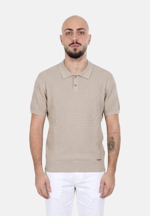Uomo calvo con barba che indossa una polo beige a maniche corte dalla trama ruvida e pantaloni bianchi, in piedi su uno sfondo chiaro uniforme.