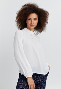 Hvid bluse med lange ærmer i let stof, med plisseret front, høj krave og elastiske manchetter. Båret med mønstrede mørke bukser.