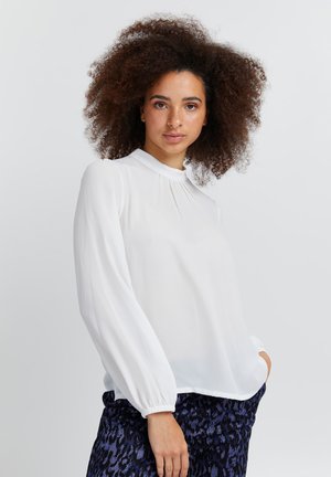 Weiße Bluse mit langen Ärmeln aus leichtem Stoff, mit einer gefältelten Vorderseite, hohem Kragen und gerafften Manschetten. Getragen mit gemusterten dunklen Hosen.