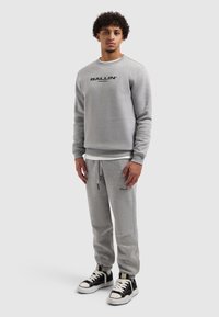Grijs sweatshirts en joggingbroek set. Het sweatshirt heeft een zwarte "BALLIN'"-logo; de broek heeft een trekkoord en subtiele merkdetails. Gedragen met zwarte sneakers.