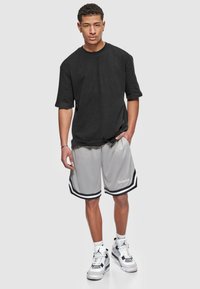 T-shirt noir oversize, shorts de sport gris avec des rayures noires et un logo, chaussettes blanches et baskets noires et blanches sur un fond simple.