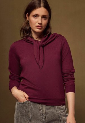 Kvinde med skulderlangt brunt hår iført guldringformede øreringe og en vinrød sweater med bundet krave og grå stribede bukser.