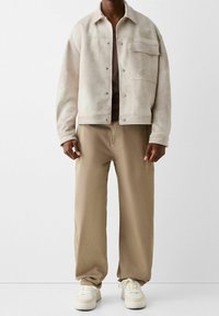 Beige mokkanahkatakki paitakauluksella ja nepparikiinnityksellä, yhdistetty vaaleanruskeisiin leveälahkeisiin housuihin ja valkoisiin tennareihin.