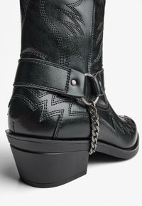 Črne usnjene kavbojske škornje z ostrim nosom, dekorativnim šivom, okrasjem iz kovinske verige in nizkim blokastim peto.