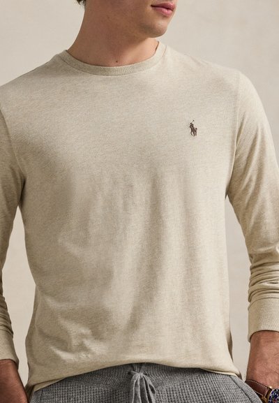 Man wearing beige long-sleeve shirt with a small dark horse and rider logo on the left chest, paired with gray knit pants.