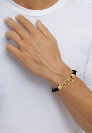 Männliches Handgelenk mit schwarzem geflochtenem Armband mit goldenem Verschluss, Finger liegen auf der Brust über einem weißen Hemd.