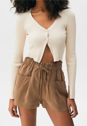 Mujer con cárdigan cropped de manga larga acanalado color crema y pantalones cortos marrones de cintura alta con cordón y bolsillos.