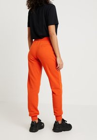 Oranžové tepláky s elastickým pasem a žebrovanými manžetami, se zadní kapsou. V kombinaci s černým tričkem s krátkým rukávem a černými teniskami.