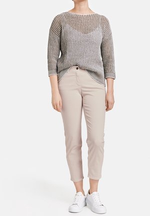 Femme vêtue d'un pull en maille gris, d'un pantalon beige raccourci et de baskets blanches, debout sur un fond blanc uni.