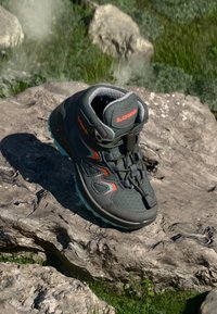 Lowa Wanderschuh in Dunkelgrün mit orangefarbenen Akzenten, mit einem hohen Knödesdesign, strukturiertem Obermaterial und gepolsteter Sohle, die auf einem Felsen ruht.