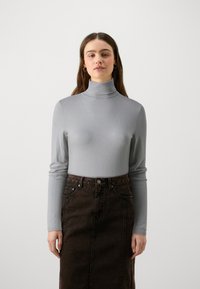 Pull en col montant gris avec de longues manches, en matière texturée, associé à une jupe en denim brun foncé avec poches avant et fermeture à boutons.