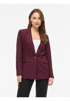 Kvinne med langt brunt hår iført en knapperød burgunderblazer over en hvit topp og svarte bukser, stående mot en ensfarget hvit bakgrunn.