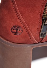 Rode suède enkelboot met een bruine chunky hak, voorzien van een metalen rits en een reliëf boomlogo aan de zijkant.