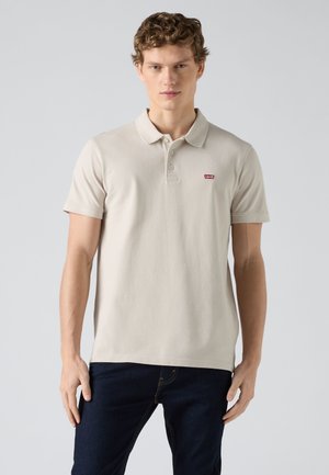 Noormees kannab beežit lühikeste varrukatega polosärki, millel on punane Levi's logo, ning tumedaid siniseid teksapükse, seisab ühevärvilise halli tausta ees.