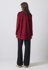 Una donna dai capelli castani mossi si trova di spalle, indossando una camicia rossa a maniche lunghe, pantaloni neri a gamba larga e scarpe beige su uno sfondo bianco.