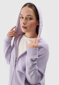 Levanduľová mikina na zips s predným vreckom, rebrovanými manžetami a kapucňou so šnúrkou, nosená cez svetlý krémový top. Hladká textúra látky.