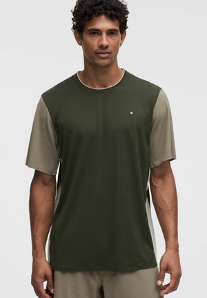 Mann trägt grünes und beiges kurzärmeliges Sportshirt mit kleinem Logo auf der Brust, steht vor hellem Hintergrund.
